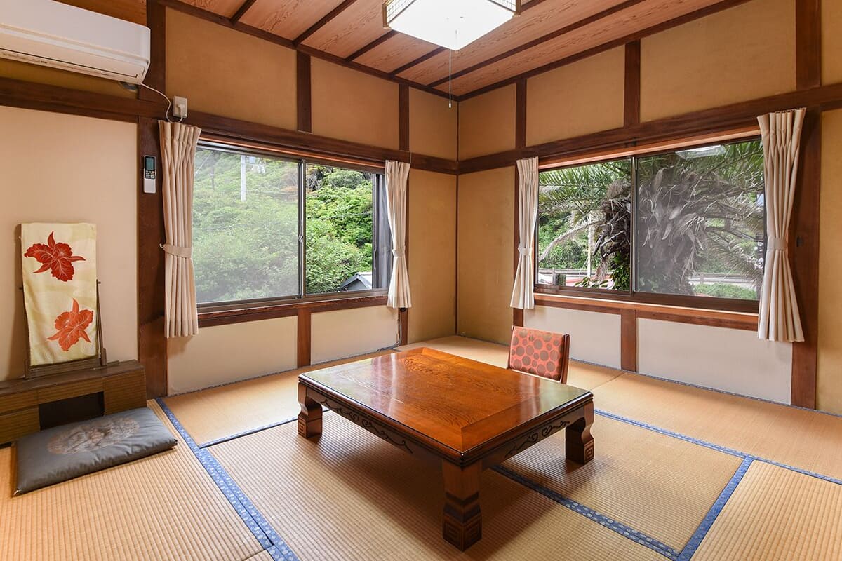 A traditional Japanese room with tatami flooring, a wooden table, and two windows with curtains.