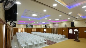 A conference room with rows of white chairs, a podium, and ceiling lights. - JR Stays (Coimbatore)