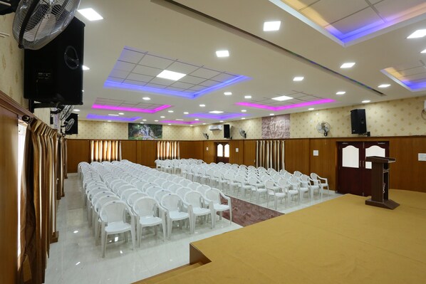 A conference room with rows of white chairs, a podium, and ceiling lights. - JR Stays (Coimbatore)