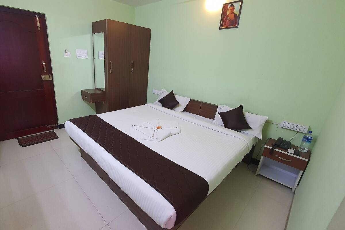 A hotel room with a bed, a wardrobe, a bedside table with a phone and water bottle, and a door.