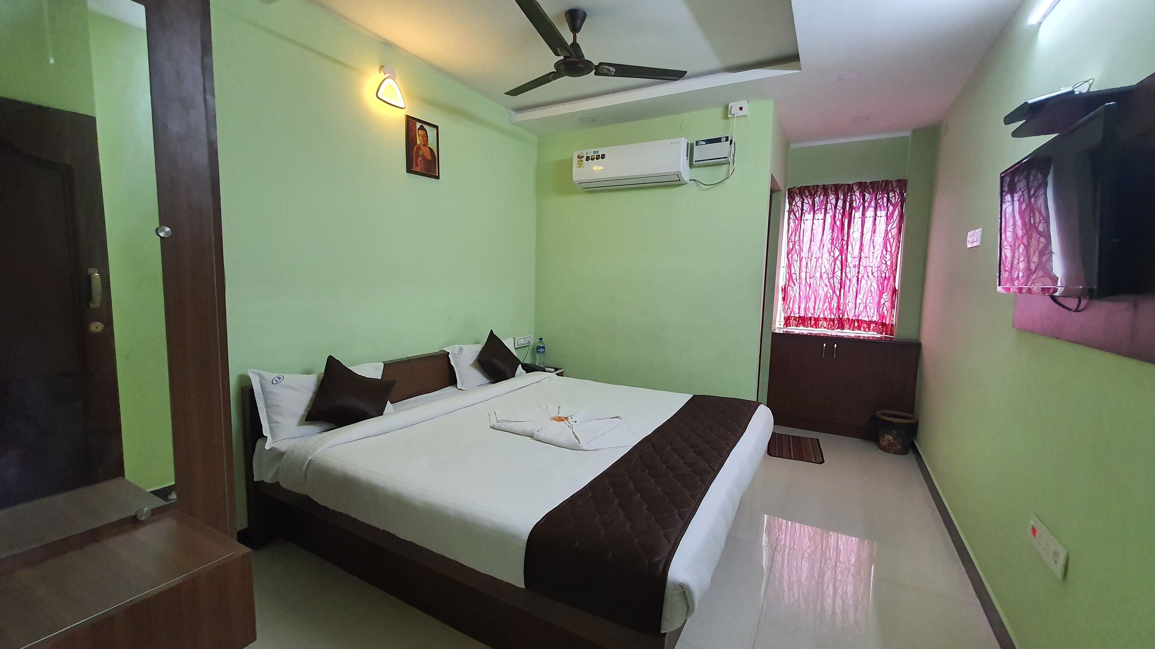 Habitación de hotel con cama, ventilador de techo, equipo de aire acondicionado, ventana con cortinas y televisor en la pared.