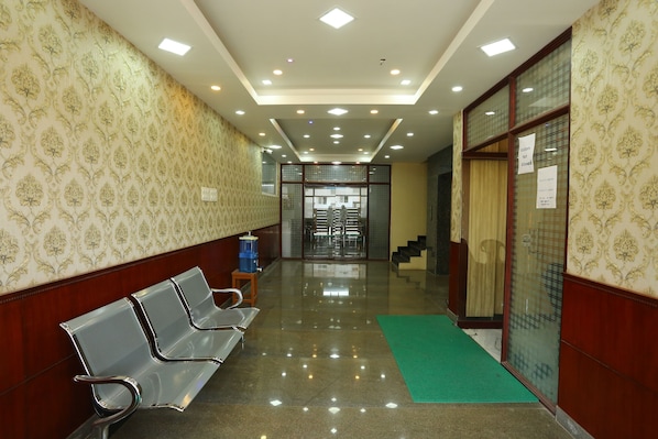 A hotel corridor with patterned wallpaper, metal chairs, a green mat, and a glass door with a sign. - JR Stays (Coimbatore)