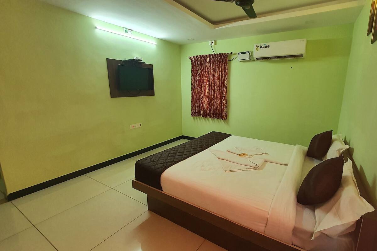 A hotel room with a bed, a flat-screen TV mounted on the wall, an air conditioning unit, and a curtain.
