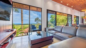 Un salon moderne avec un grand canapé, une table basse et un téléviseur. De grandes fenêtres offrent une vue sur l’extérieur.