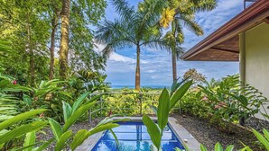 Une piscine entourée de plantes tropicales et de palmiers, avec vue sur l’océan.