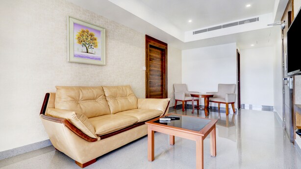 A living room with a beige leather sofa, a glass coffee table, and two armchairs. There is a wooden door and a framed picture on the wall. - Treebo Trend Royal Park (Thiruvananthapuram)