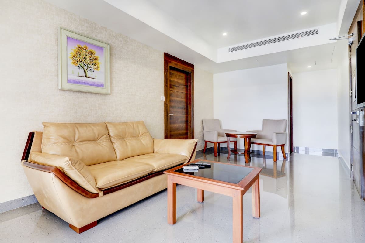 Una sala de estar con un sofá de cuero color beige, una mesa de centro de vidrio y dos sillones. Hay una puerta de madera y un cuadro enmarcado en la pared.