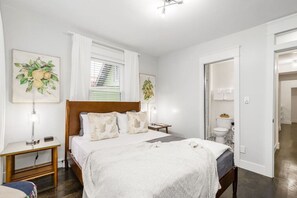 A bedroom with a wooden bed, white bedding, a bedside table with a lamp, and a window with blinds. - 5 Bedroom Group Stay | Private Bath Every Room (KCMO)
