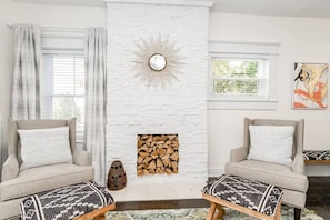 A living room with a white brick fireplace, two armchairs, a patterned rug, and a painting on the wall.