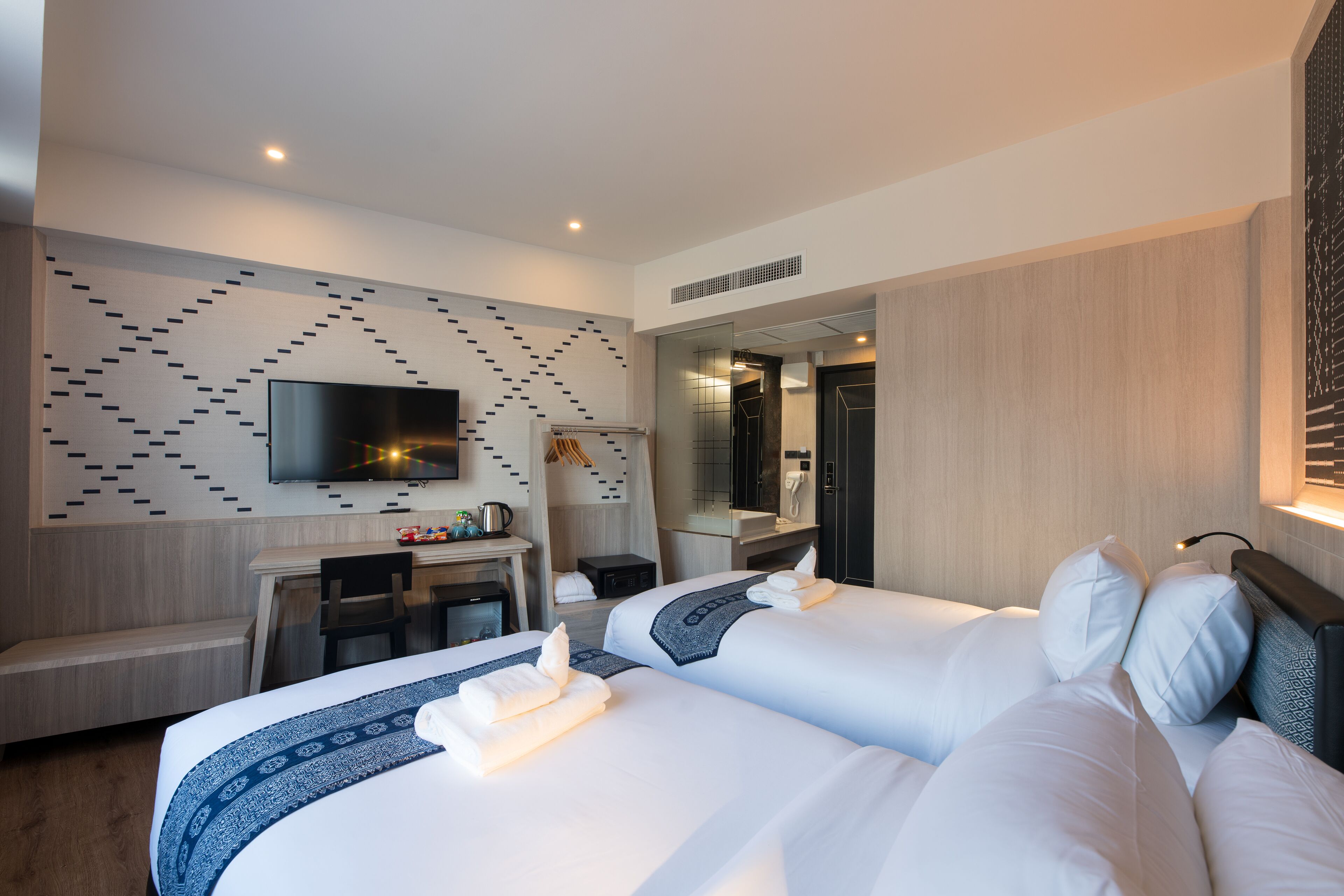 A modern hotel room with two beds, a desk, a flat-screen TV, and a bathroom visible through a glass partition.