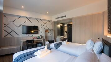 A modern hotel room with two beds, a desk, a flat-screen TV, and a bathroom visible through a glass partition.