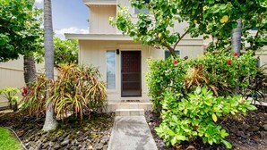 Un chemin menant à un bâtiment doté d’une porte, entouré de plantes tropicales et d’un petit jardin.