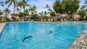 Une piscine à l’eau d’un bleu cristallin, entourée de chaises longues et de parasols, avec des palmiers et des bâtiments en arrière-plan.