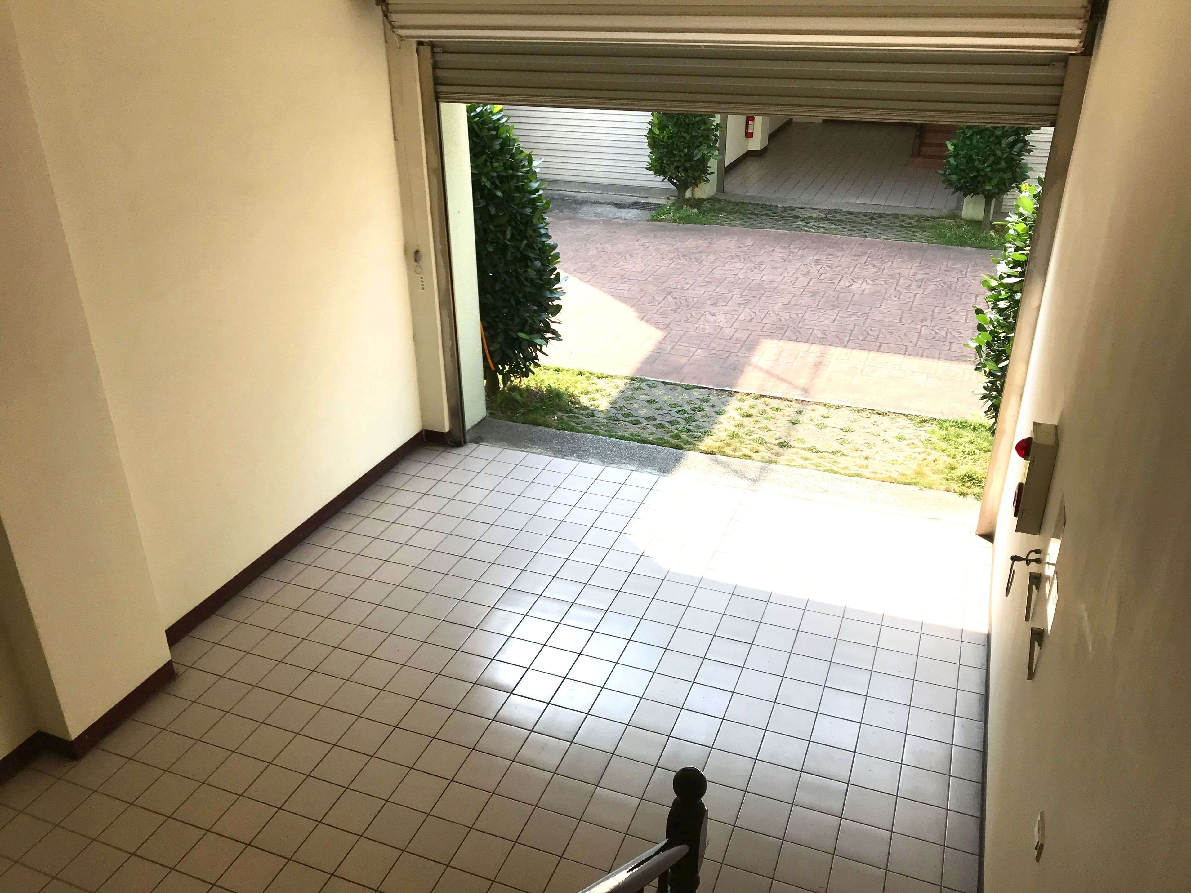 A tiled floor leading to a garage door with a view of a paved driveway and small trees.