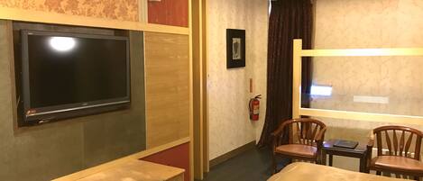 Habitación de hotel con televisión de pantalla plana en la pared, un banco de madera, una cama con cubierta color beige, una mesita con una silla y un extintor en la pared.