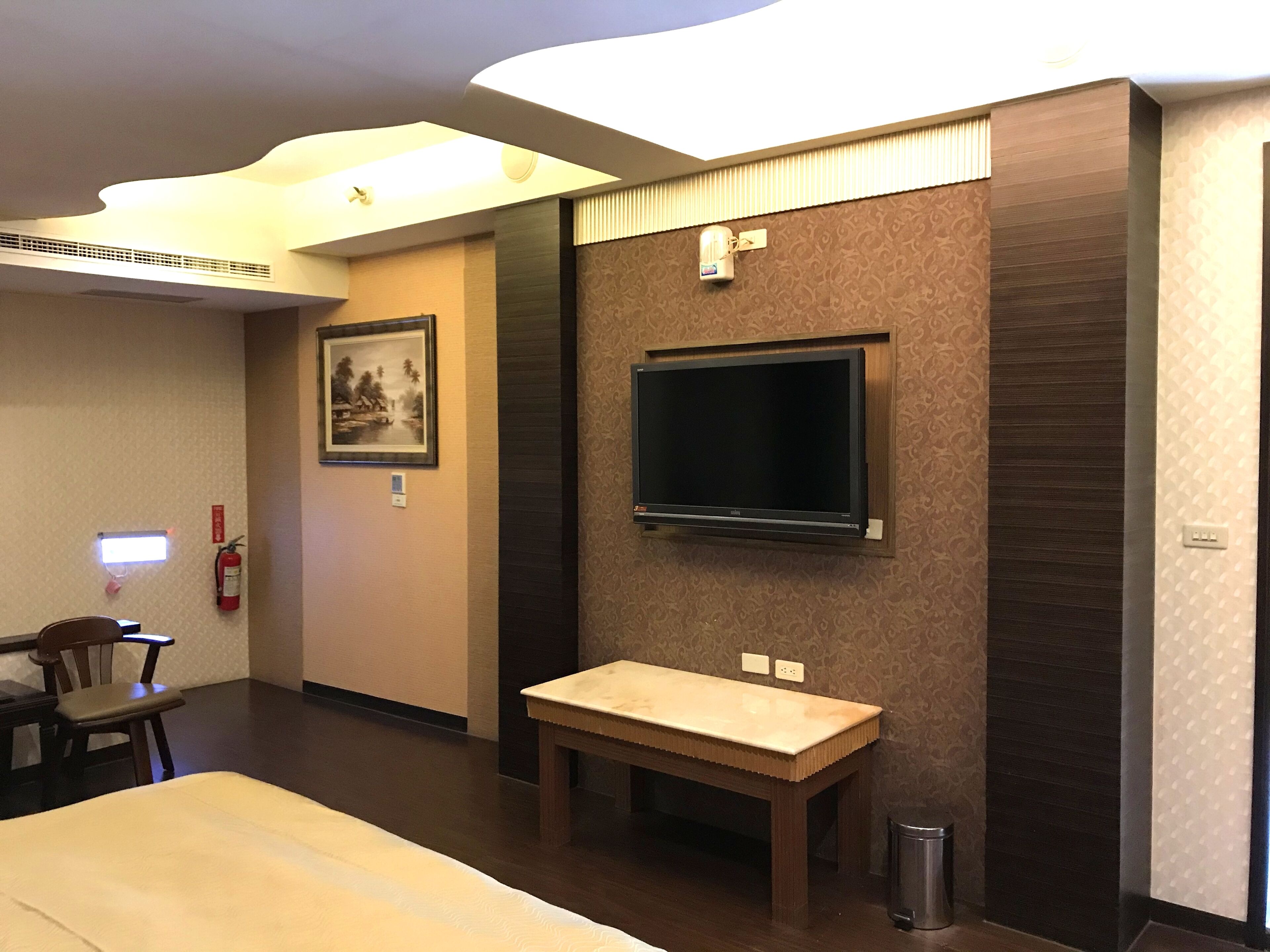 Hotellihuone, jossa on sänky, pieni puinen pöytä, seinälle asennettu televisio, kehystetty kuva ja palosammutin.