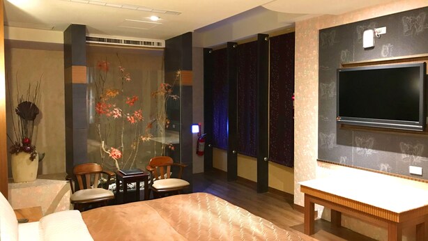 A modern hotel room with a large bed, a desk, a flat-screen TV mounted on the wall, and a decorative plant arrangement. - Mapleton Motel (Taichung)