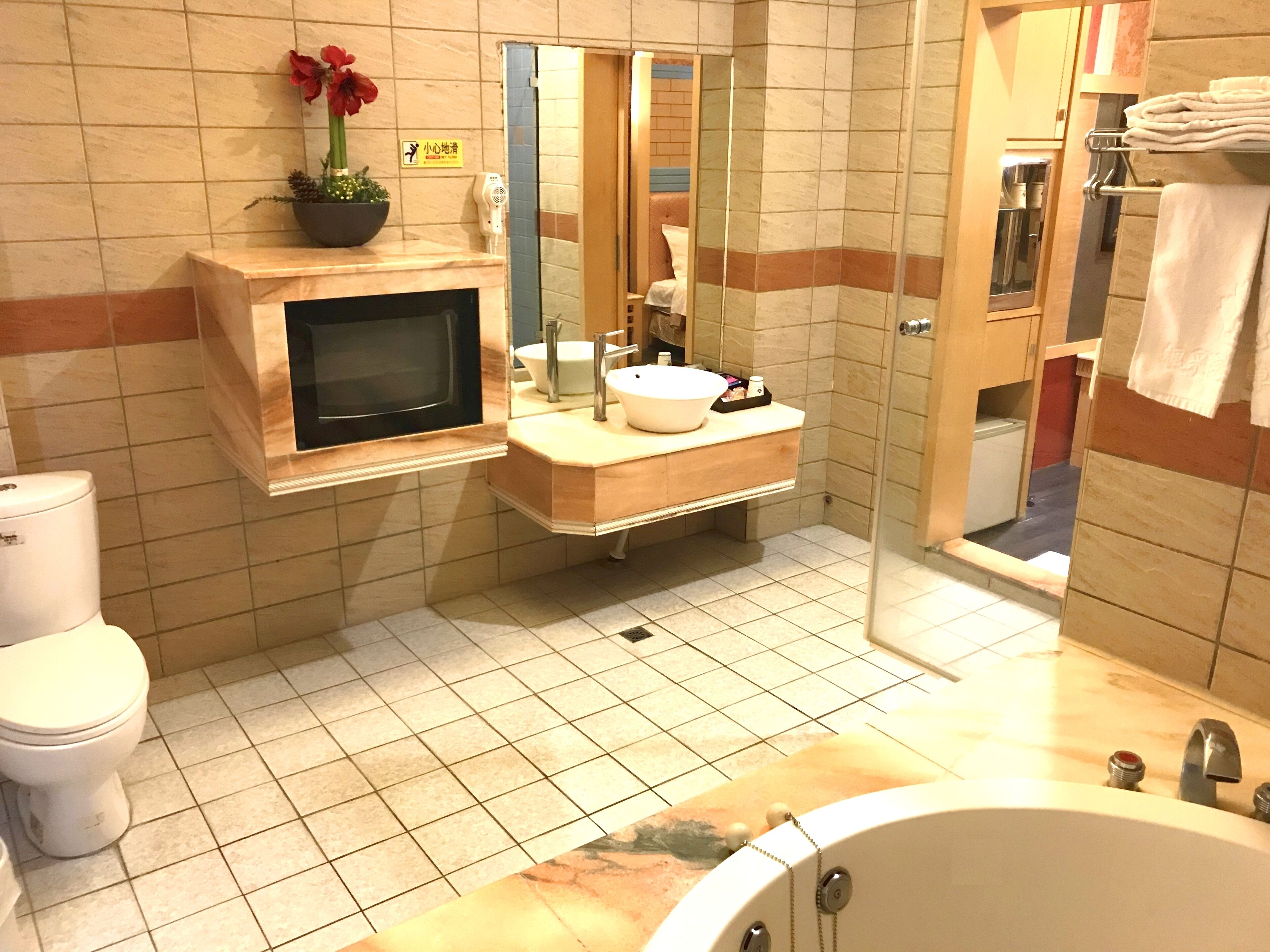 Un baño con inodoro, lavamanos y bañera. Hay un televisor montado en la pared y una planta en maceta sobre el mostrador.