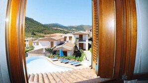 Vista dalla finestra su una zona piscina con lettini, un piccolo edificio e un paesaggio collinare sullo sfondo.