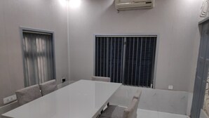 A conference room with a large white table, four grey upholstered chairs, a window with blinds, an air conditioning unit, and a door. - Maddy's Den for females (Kolkata)