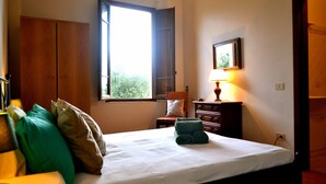 Ein Schlafzimmer mit Bett, einem Fenster mit geöffneten Lamellen, einem Schminktisch, einer Lampe und einem gerahmten Bild an der Wand.