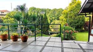 Um terraço com plantas em vasos, uma mesa estampada e vista para um jardim exuberante.
