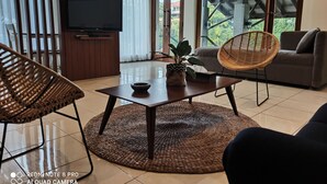 Uma sala de estar moderna com uma televisão grande, cadeiras de vime, um sofá e vista para o verde lá fora.