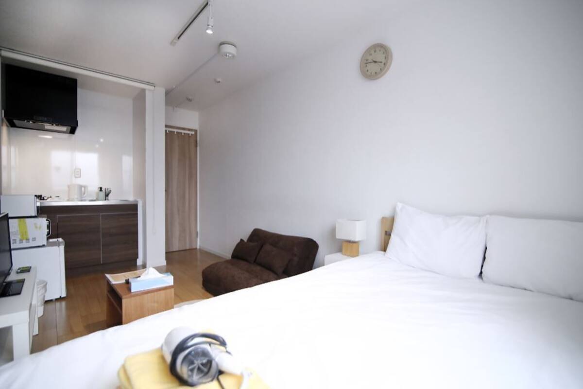 Ein modernes Hotelzimmer mit einem Bett, einem Schreibtisch mit Computer, einer kleinen Küchenzeile und einem braunen Sessel.