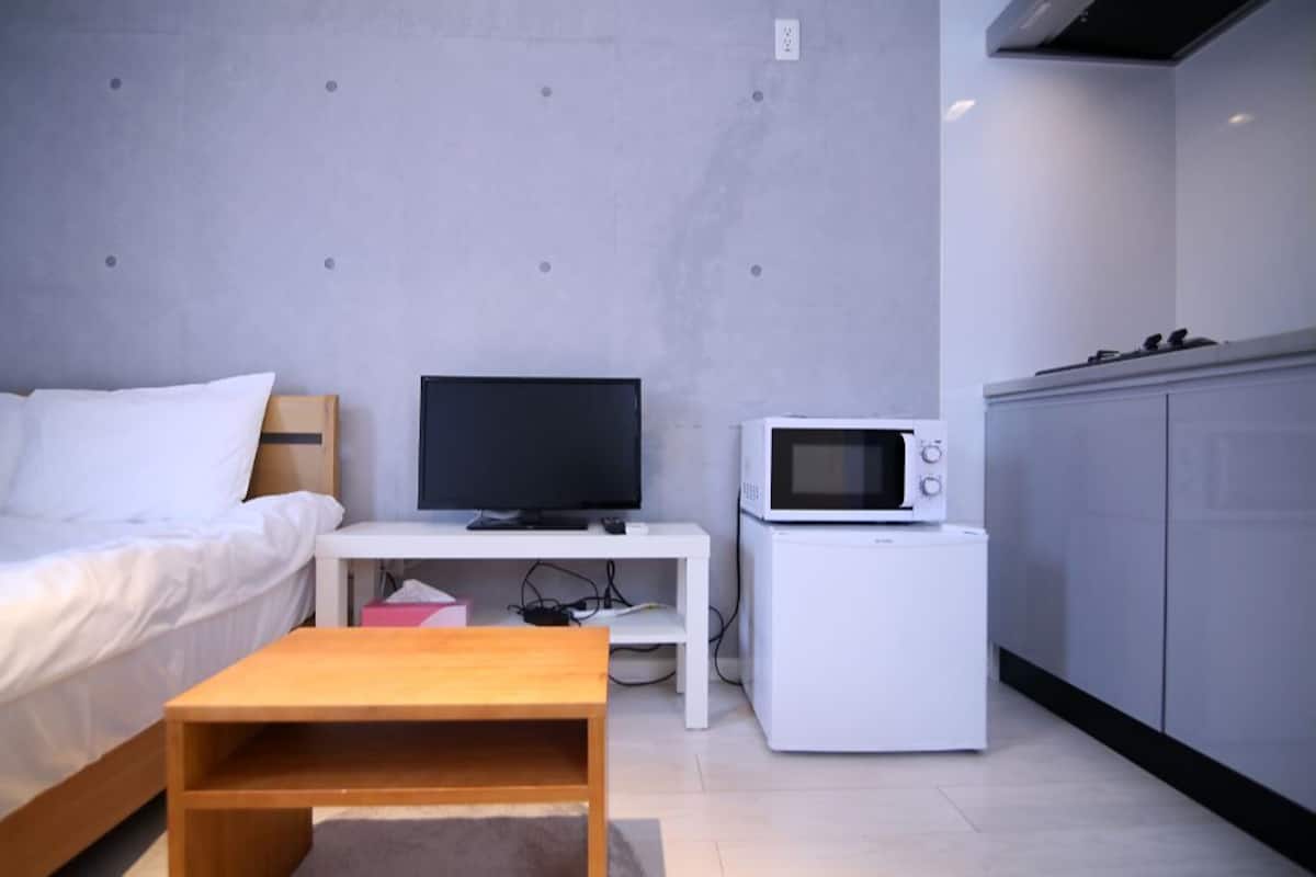Ein modernes Zimmer mit einem Bett, einem Fernseher auf einem weißen Standfuß, einer Mikrowelle und einem kleinen Holztisch.