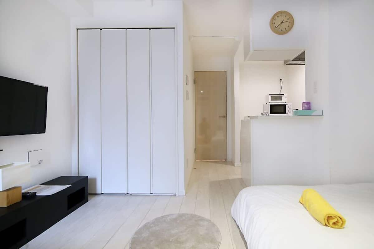 Ein Hotelzimmer mit Bett, Fernseher, Schreibtisch, Kleiderschrank und einem Badezimmer mit Spiegel. An der Wand hängt eine Uhr.