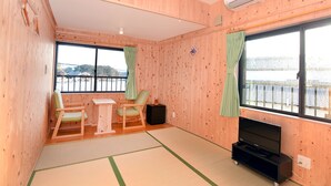 Et værelse med træpaneler, en tatami-gulvbelægning, et fjernsyn, et lille bord, to stole og udsigt til et vandareal gennem store vinduer.