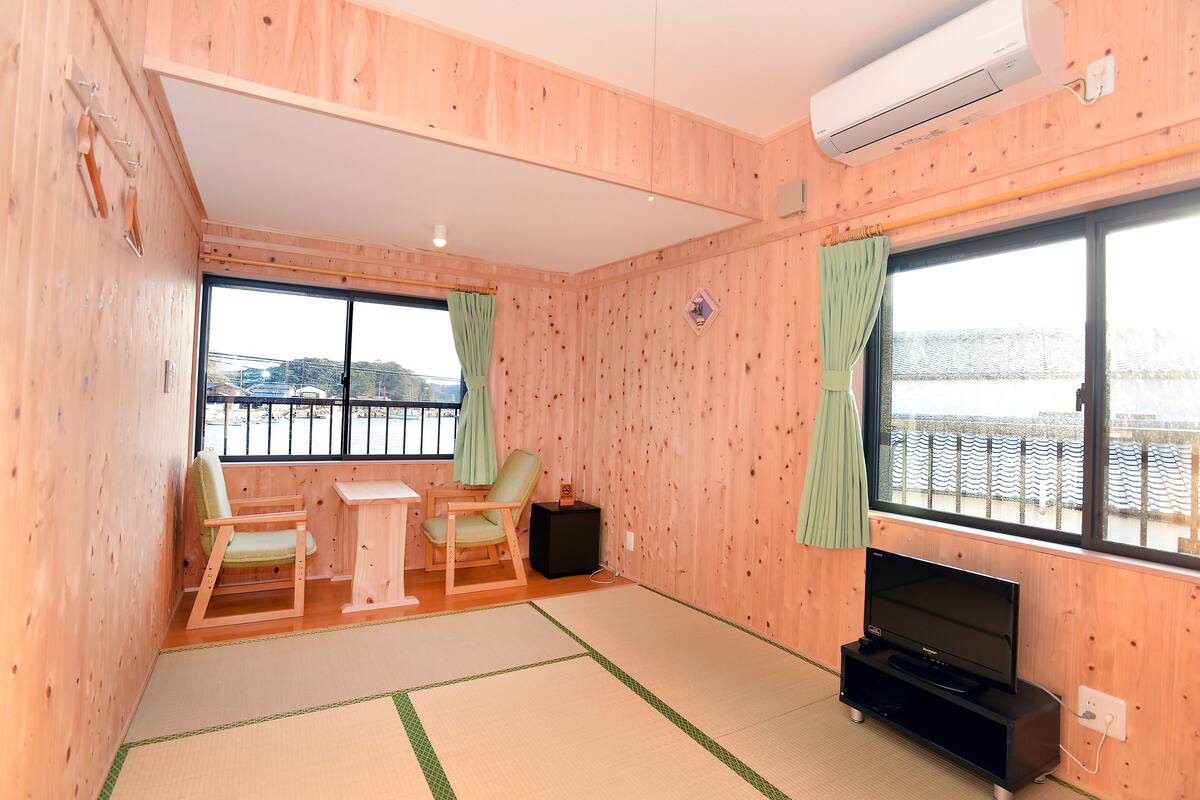 Ein Zimmer mit Holzwänden, einem Tatami-Bodenbelag, einem Fernseher, einem kleinen Tisch, zwei Stühlen und Blick auf ein Gewässer durch große Fenster.