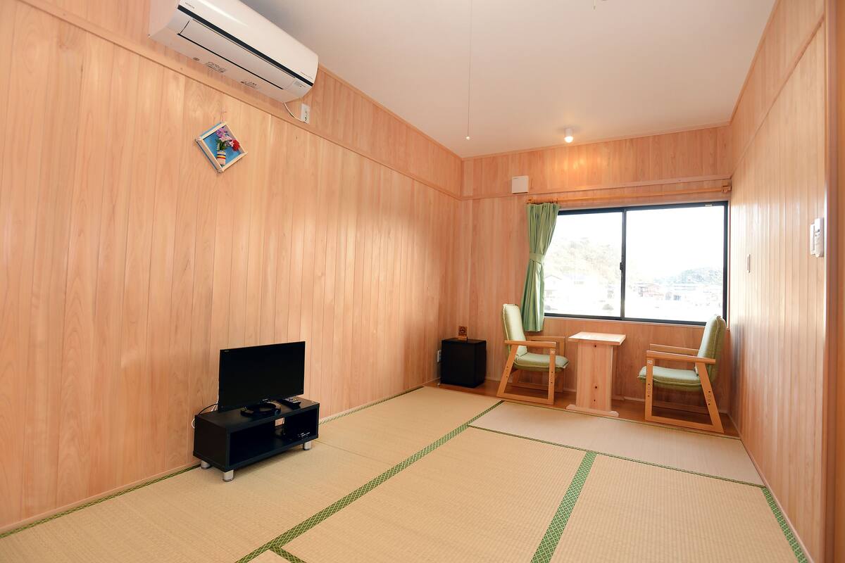 Ein Zimmer mit Holzwänden, einem Tatami-Boden, einem Fernseher, einem Lautsprecher und einem Fenster mit Vorhang.