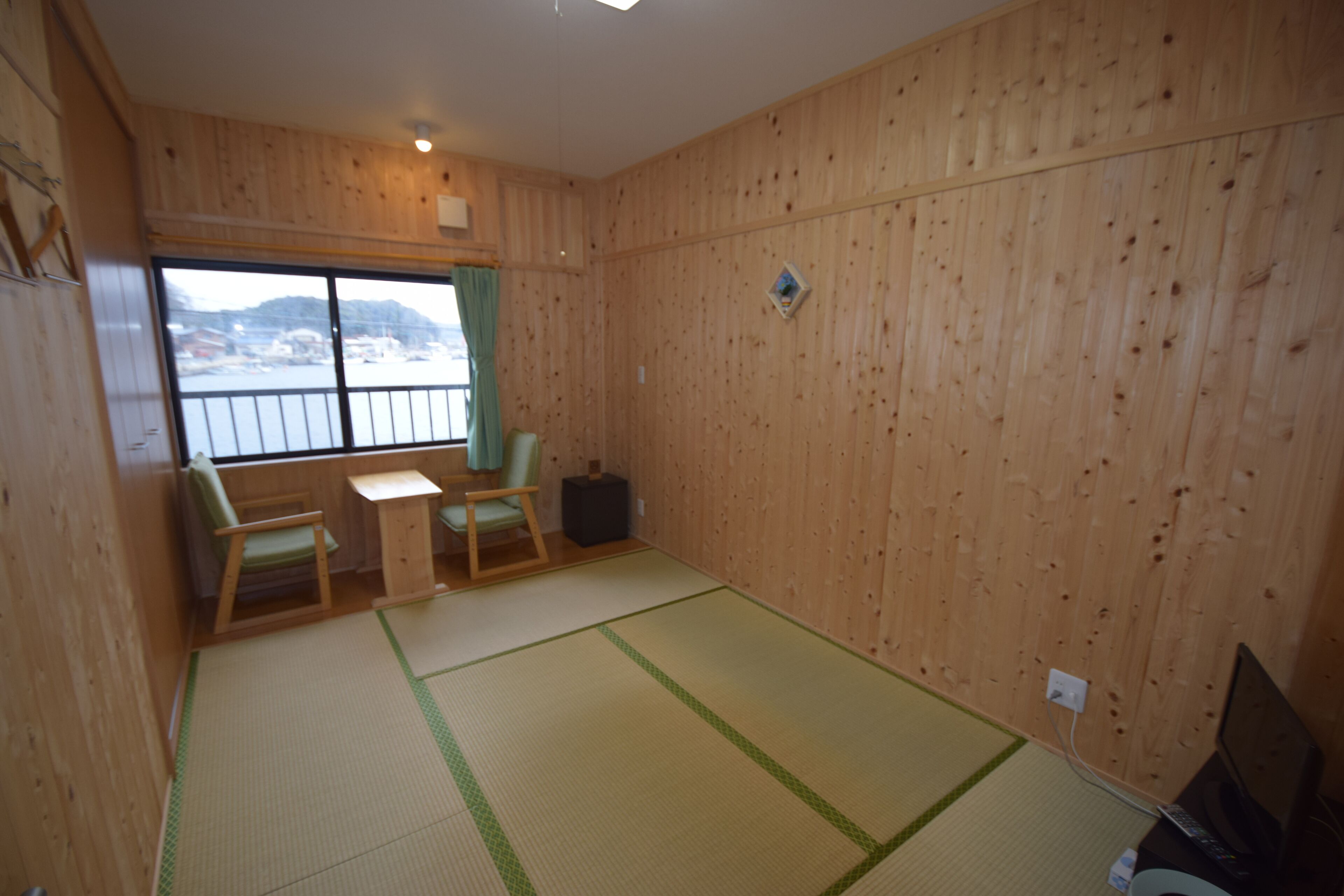 Een kamer met houten wanden, tatami matten, een kleine tafel, twee stoelen, een televisie en een raam met uitzicht op een waterpartij en heuvels.