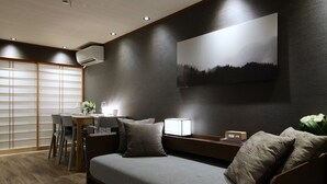 A modern hotel room with a sofa, a desk with chairs, a wall-mounted air conditioner, and a painting of a mountain landscape. - Shiki Homes IKKŌ 8 (Kyoto)