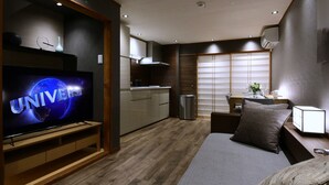 A modern hotel room with a TV displaying 'UNIVERSAL', a wooden floor, a bed with a pillow, a bedside table with a lamp, and a view of a balcony with a window. - Shiki Homes IKKŌ 8 (Kyoto)