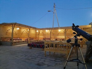 Ein Sitzbereich im Freien mit Holzbänken und -tischen, ein Teleskop auf einem Stativ und ein Holzgebäude mit Rundfenstern im Hintergrund.