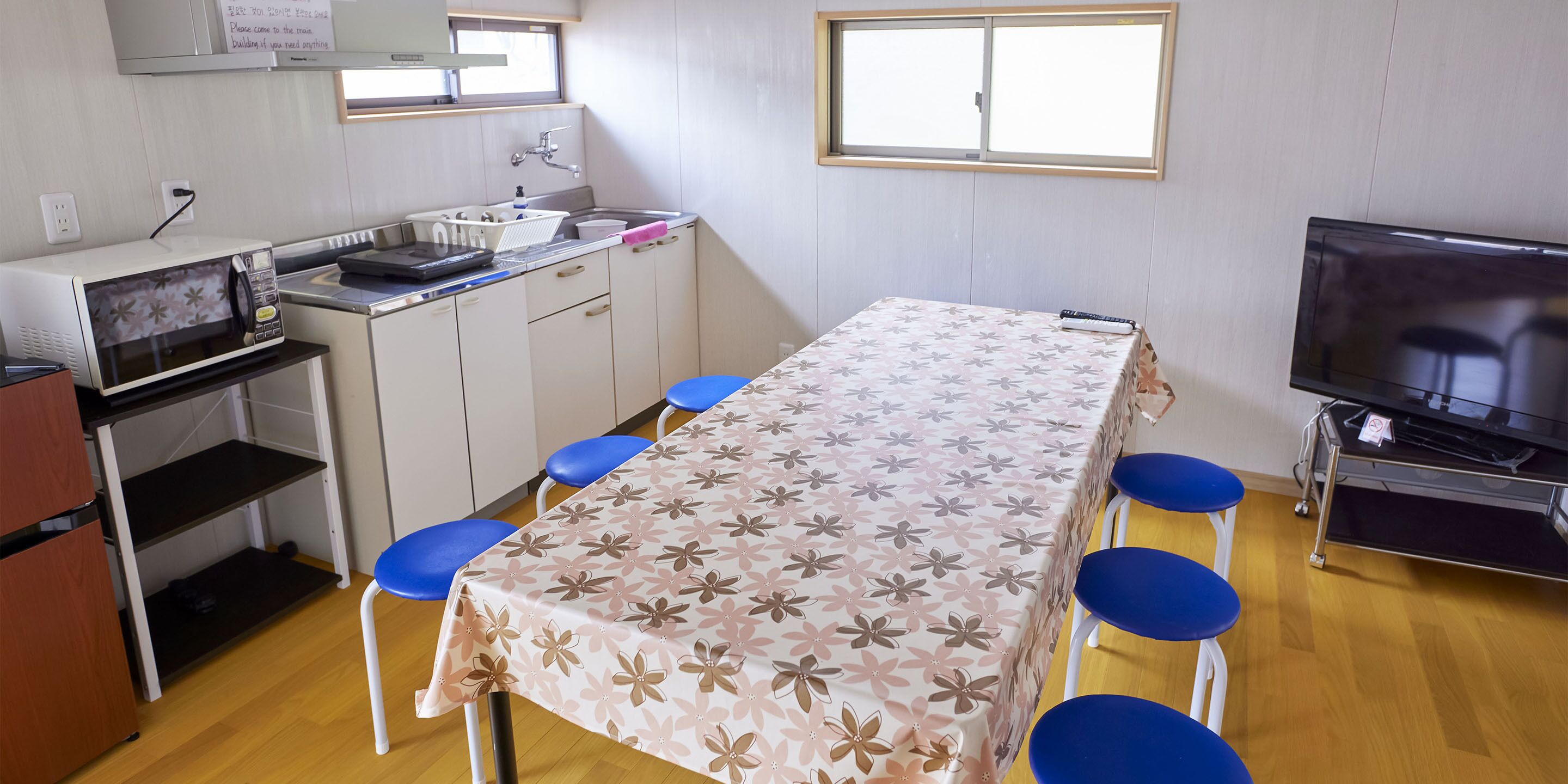 A compact room with a kitchenette, a dining table with blue chairs, a television, and a window providing natural light.