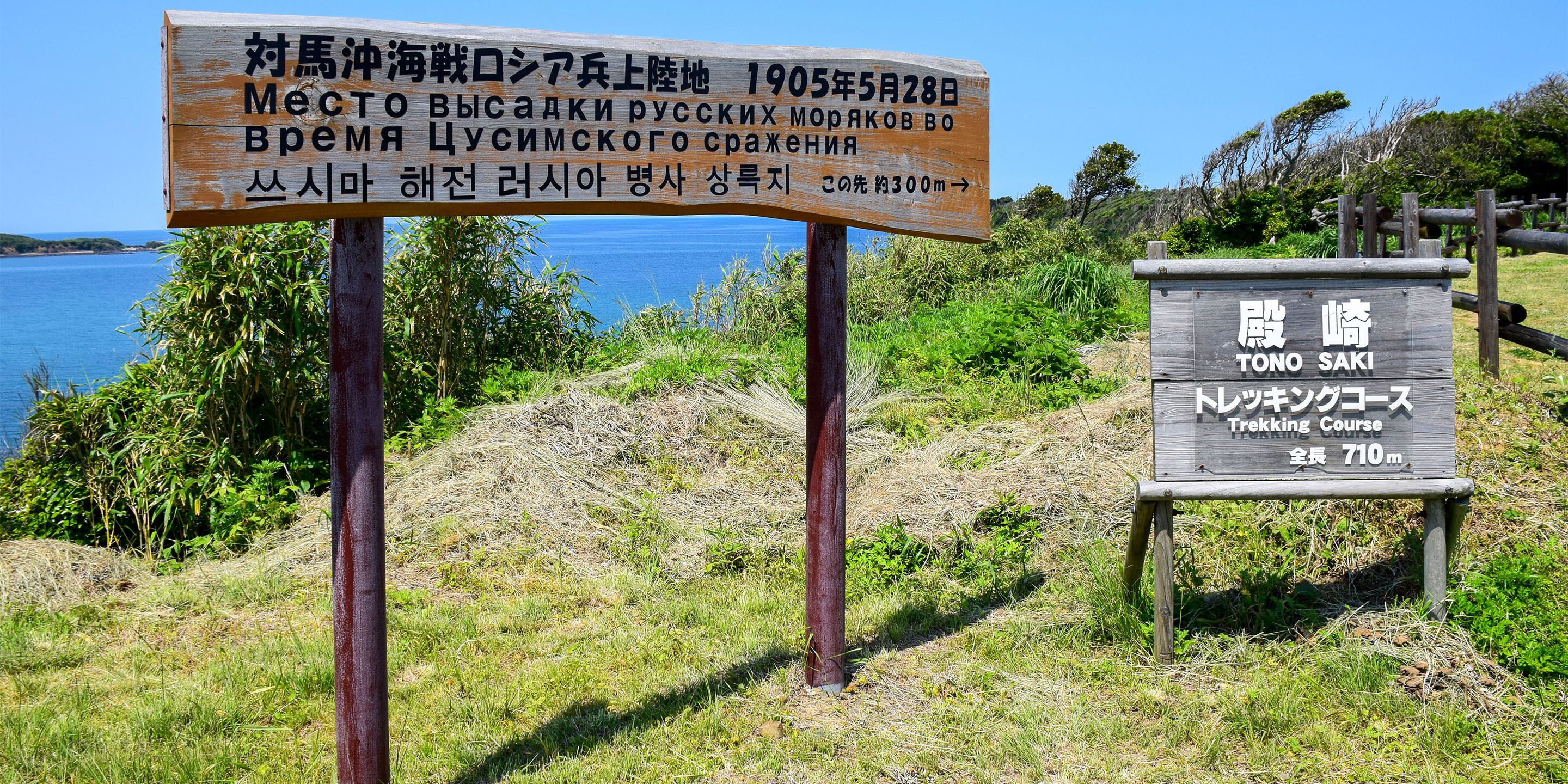 A wooden sign with text in multiple languages, a smaller sign indicating a trekking course, and a natural landscape with water and greenery in the background.