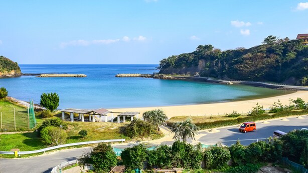 A coastal area with a sandy beach, a road with a van, and a building on a hill. - Hanareya (Tsushima)