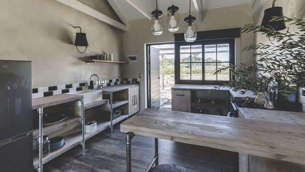 Una cucina con un tavolo di legno, lampade a sospensione e vista sull'esterno attraverso una grande finestra.
