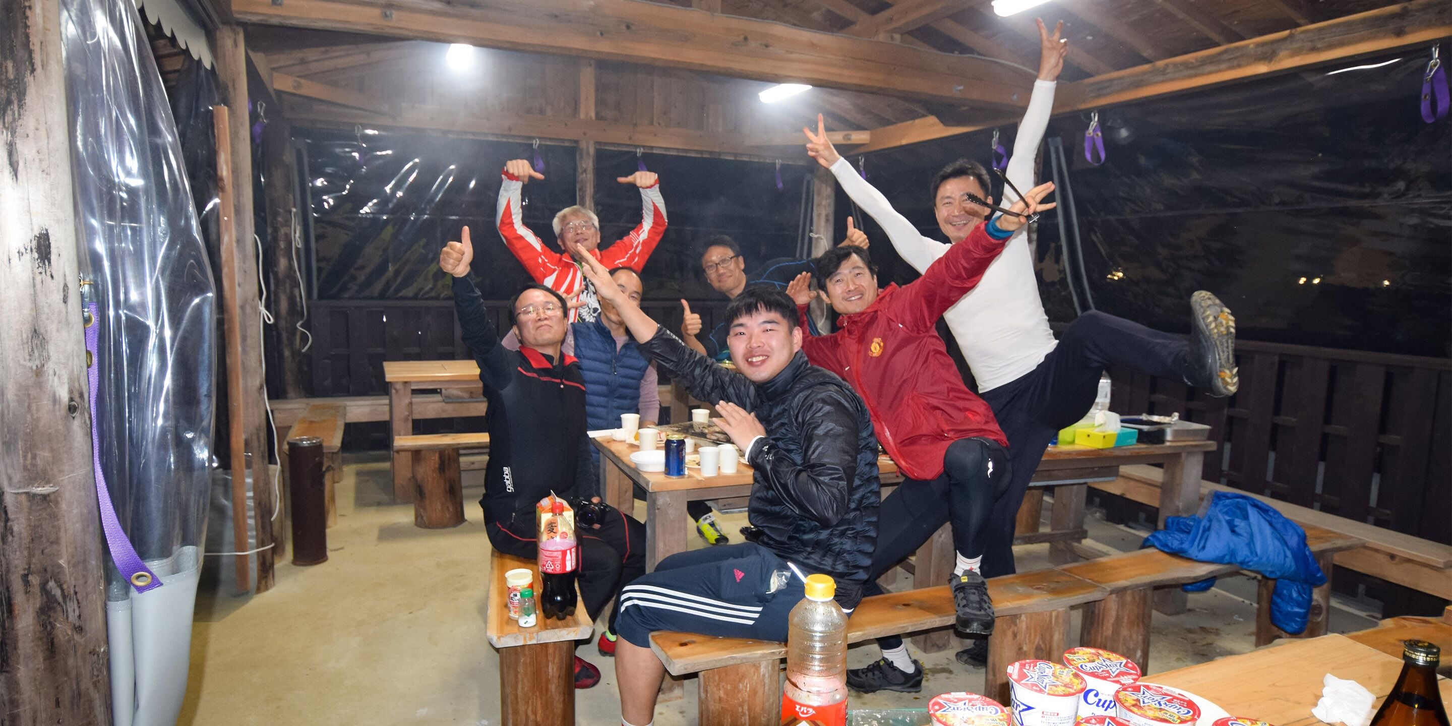 A group of people in a wooden cabin with tables and benches, some holding drinks and snacks.
