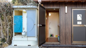 Une unité de toilettes portative portant le numéro 48741, une porte en bois avec une pancarte indiquant « Salle de douche n° 2 », et une petite étagère contenant des produits de nettoyage.