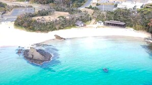 Vue aérienne d’une zone côtière avec des eaux turquoise limpides, une plage de sable et quelques bâtiments, dont une maison et une construction dotée d’un toit.