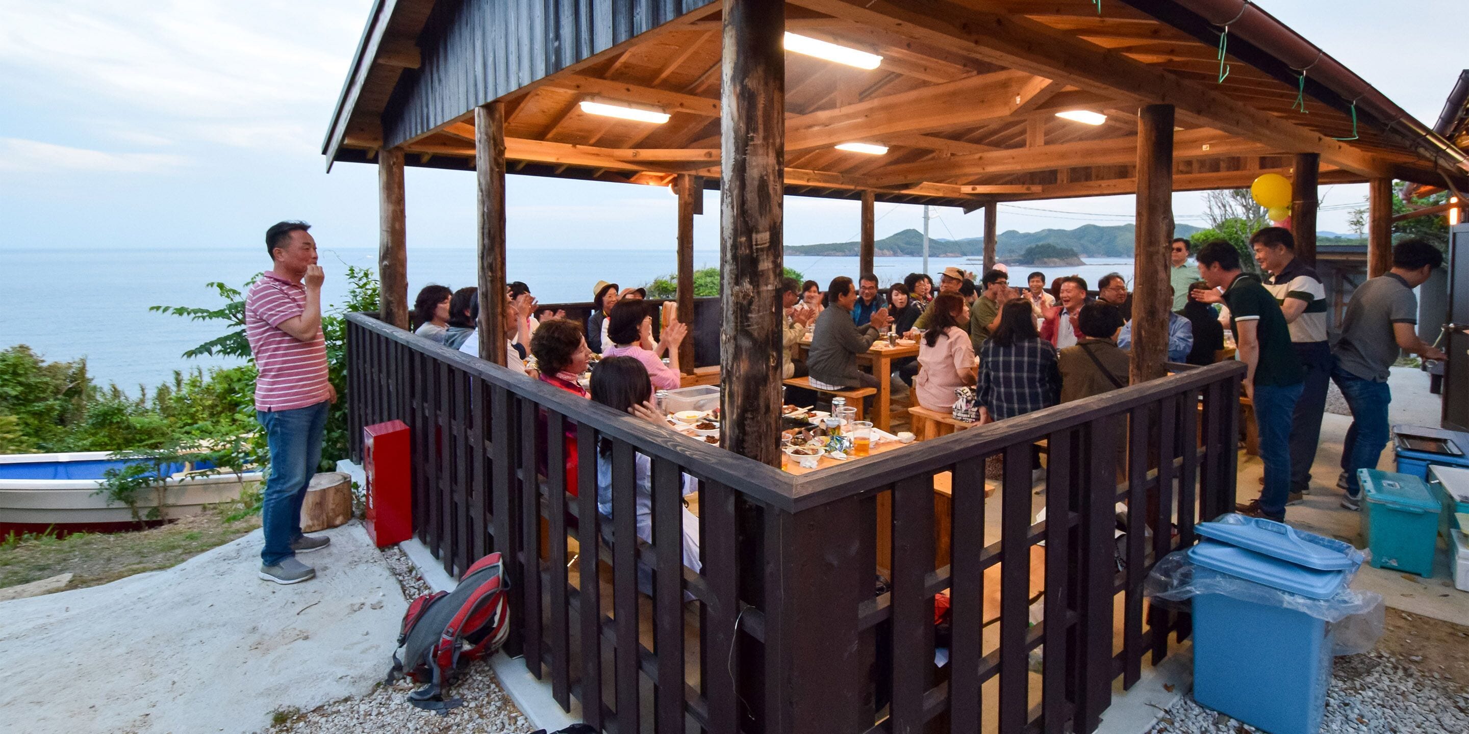 Un espacio comedor al aire libre con techo y vistas al mar, mesas de madera y un grupo de personas cenando.