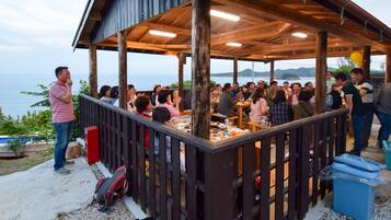 Un espacio comedor al aire libre con techo y vistas al mar, mesas de madera y un grupo de personas cenando.