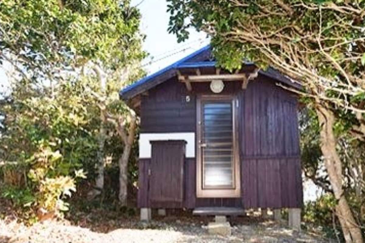 Eine kleine Holzhütte mit dunklem Dach, einer Tür und einem Fenster, umgeben von Bäumen und Grün.