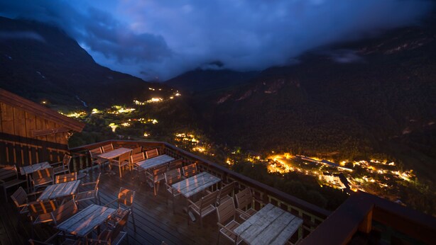 A wooden deck with tables and chairs overlooking a valley with illuminated buildings. - Westerås Gard (Stranda)