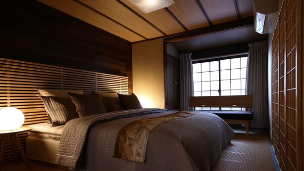 Ein traditionelles japanisches Schlafzimmer mit Tatami-Boden, einem niedrigen hölzernen Bett, einem Nachttisch mit Lampe und Blick ins Freie durch einen Shoji-Schirm.