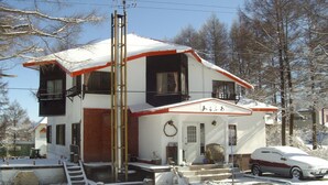 A two-story building with a red and white exterior, a visible front door, and a car parked in front. - Alpha Lodge Madarao (Myoko)